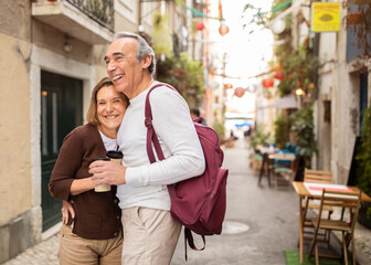 Joyful Senior Tourists Couple Hugging Posing With Backpack Holding Paper Coffee Cups On Cozy Lisbon...