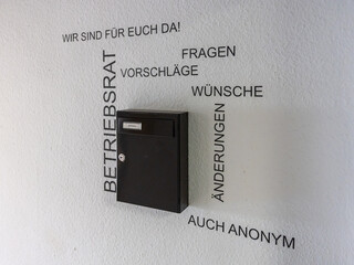 Das Bild zeigt einen schwarzen Briefkasten an einer Wand.Um den Briefkasten sind Begriffe wie „Betriebsrat“, „Fragen“, „Vorschläge“, „Wünsche“, „Änderungen“ und „Auch anonym“ auf die Wand gedruckt.