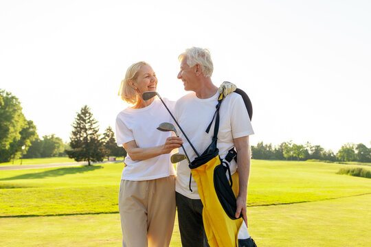happy elderly senior couple in white uniform holding golf clubs and hugging on golf course at sunset, old man and woman playing golf and smiling looking at camera in nature