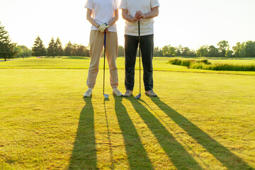 Fototapeta premium man and woman playing golf on the course at sunset, close-up of golf ball and clubs and people's feet on green grass, couple competing in the game and hitting the ball into the hole