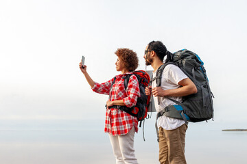 young african american couple tourists with hiking equipment and backpacks standing near the sea and taking selfie on smartphone, man and woman traveling with tents and photographing horizon on mobile