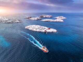 Aerial view of floating fishing boat in blue sea in snowy Henningsvaer village, Lofoten islands,...