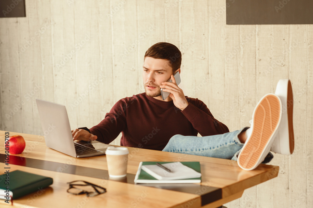 Poster freelancer guy talking on mobile phone working on laptop computer sitting with legs on desk in offic