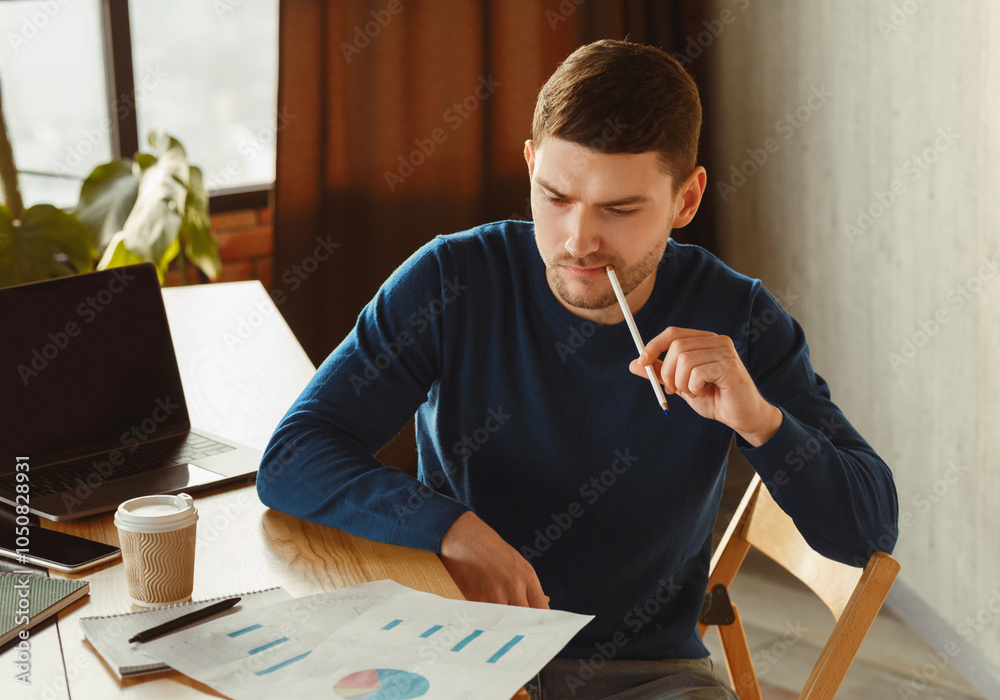 Poster thoughtful millennial guy looking through data graphs and charts thinking on business startup sittin