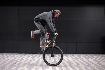 Naklejka premium young hipster guy doing stunts on a bike in the city against the background of a black wall, street rider shows freestyle on a bike and makes dangerous moves