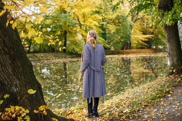 Woman in houndstooth coat stands by lake in public park. Autumn fashion clothing