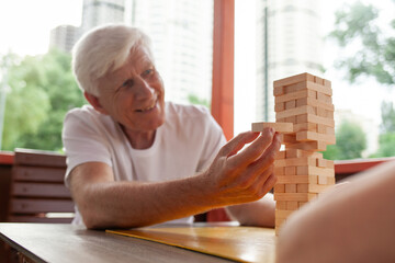 elderly couple of seniors in white t-shirts playing board game jenga in park outdoors, old man and woman building a tower of wooden bricks and competing in a game of balance