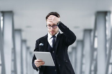 Bad News. Confused and lost adult manager in glasses using digital tablet, touching his head