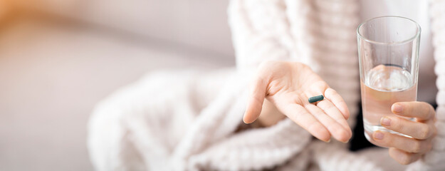 Naklejka premium Close up of sick woman taking treatment at home, holding glass of water and pill