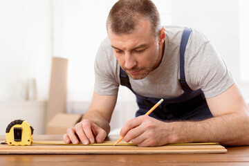 Carpenter At Work. Mature Craftsman Working Making Furniture Measuring Wooden Board Indoors, Wearing Blue Coverall Uniform. Carpentry Profession And Service. Selective Focus