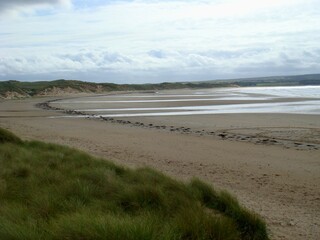 Beautiful sandy beach in Thurso, Caithness, Scotland, with grassy dunes and wide, open coastal views. Footprints lead across the sand, and the waves gently wash ashore under a partly cloudy sky.