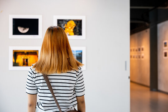 Asian woman standing she looking art gallery in front of colorful framed paintings pictures on white wall, people watch at photo frame to leaning against at show exhibition artwork gallery, Back view