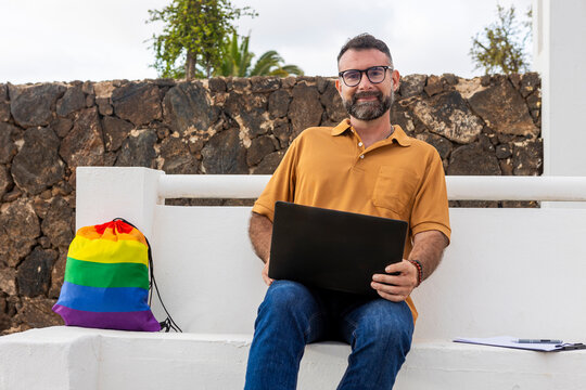 Gay Man with laptop and rainbow pride bag on white bench