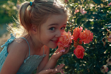 Naklejka premium A cheerful young girl with blonde hair leans closely to colorful roses in a garden, showing her delight as she enjoys the scent in the warm sunlight
