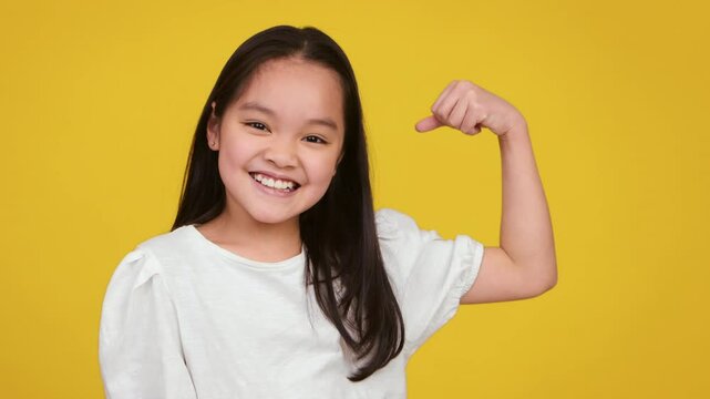A cheerful Asian girl flexes her arm muscles, displaying confidence and strength while smiling at the camera.