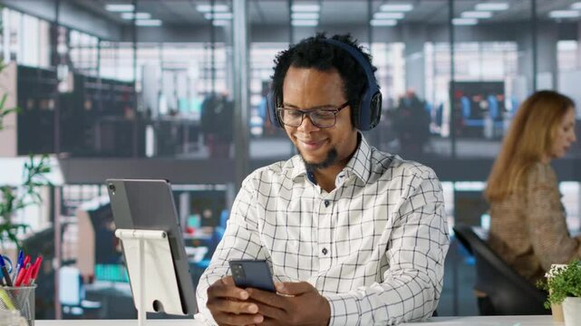 African american general manager reviews forecasts while he listens to music on headset, feeling relaxed and multitasking at his desk. Data analyst focuses on objectives, strategy planning. Camera A.