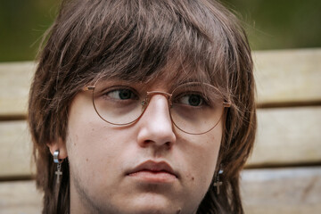 A detailed close-up of a young man wearing round glasses and a cross earring, with a serious...