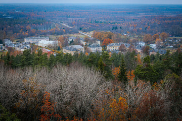 Aerial view of a small town nestled within a vast autumn forest, with trees displaying vibrant orange and yellow foliage. The town's buildings blend into the natural landscape. Zilaiskalns