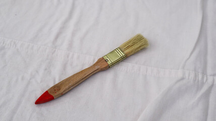 A wooden-handled paintbrush with natural bristles and a red-tipped handle lies on a white fabric background, showcasing its rustic design and practical use for painting or crafting projects.