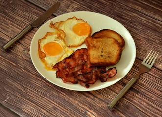 A large plate of delicious and nutritious breakfast on a wooden table, scrambled eggs, fried bacon and toasted bread, cutlery nearby.