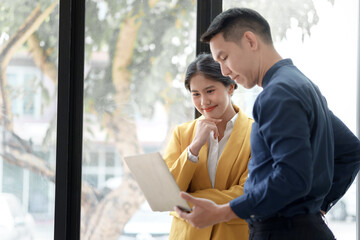 A professional woman in a yellow blazer presents a laptop to a colleague in a bright, modern office setting.