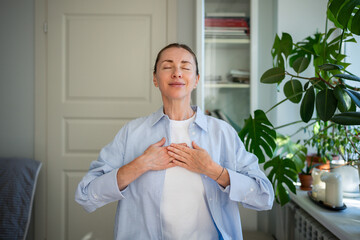 Relaxed smiling woman meditates with hands on chest, practicing breathing exercises at home....