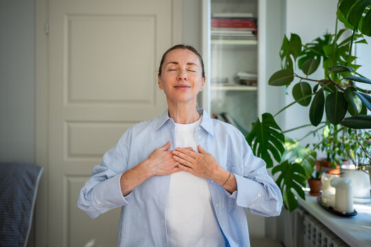 Relaxed smiling woman meditates with hands on chest, practicing breathing exercises at home. Mindfulness practice, focus on breath, self care routine, stress relief, reflection and holistic wellness