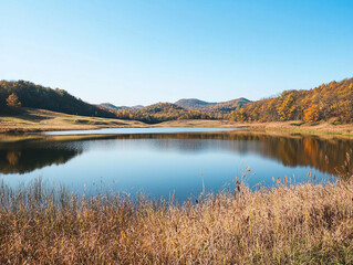 A serene lake surrounded by autumn-colored hills, with clear blue skies overhead and tall grasses on the shore