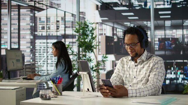 African american general manager reviews forecasts while he listens to music on headset, feeling relaxed and multitasking at his desk. Data analyst focuses on objectives, strategy planning. Camera B.