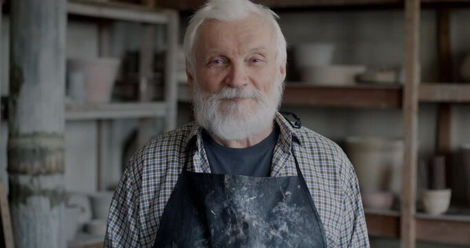 Slow motion portrait of senior man in apron standing in pottery studio smiling looking at camera. Small business and experienced artisans concept.