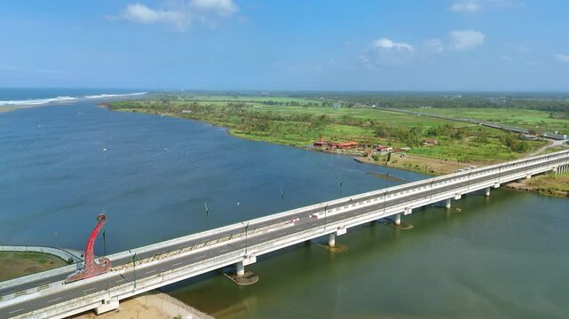Aerial view of a bridge spanning a river estuary with view of wavy coastline. Blue sky on the horizon. Kretek II Bridge, Depok Beach, Indonesia.