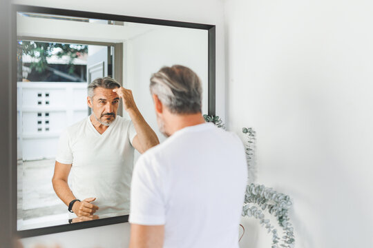 Mature man grooming in front of a mirror, adjusting hair with focused expression, wearing a white shirt - Powered by Adobe