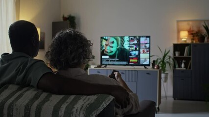 Rear shot of loving spouses sitting on sofa in modern living room and choosing movie on TV using streaming service, husband hugging wife from behind - Powered by Adobe