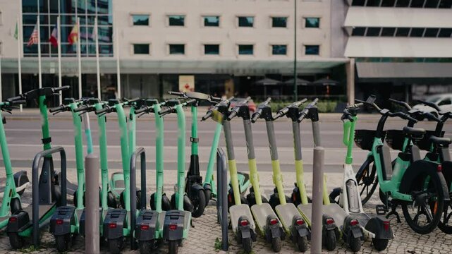 A row of green electric scooters and bikes are neatly parked on a city sidewalk, with a modern multi-story building in the background, representing sustainable urban transportation.