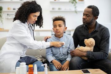 Obraz premium African female doctor listens to heartbeat of young boy while father watches closely at home. Child holds a teddy bear, symbolizing comfort during medical examination.