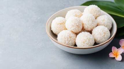 Coconut balls in a bowl, with tropical leaves and flowers on a textured surface.