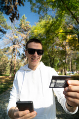 Fototapeta premium Young man online shopping using credit card and mobile phone in the park in autumn