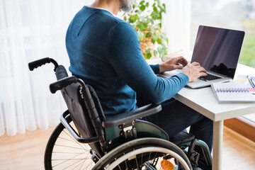 Obraz premium Close up of man in wheelchair working from home office, using computer in office. Telecommuting and home office.