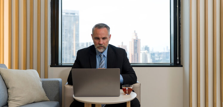 Senior businessman in suit and tie typing on laptop computer while sitting in cafeteria during coffee break. The view outside the back window is an office buildings at high-rise building district area