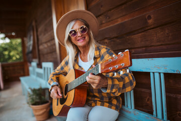 Beautiful older woman is sitting in front of her cabin, playing on guitar.