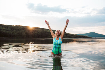Fototapeta premium Cold water swimming for elderly women. Senior sporty women standing in lake during cold evening, arms in the air.