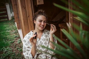 Beautiful woman doing her skin care routine outdoors, wearing bathrobe. Woman using facial skin serum with dropper.