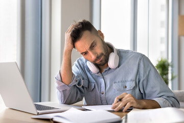 Tired focused adult Latin student man getting difficult task, sitting at work table with laptop,...