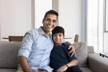Cheerful Indian dad hugging cute preteen son, sitting on home sofa, looking at camera, smiling,...