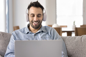Positive handsome Hispanic man in wireless headphones using laptop on couch with computer, typing, chatting online, smiling, laughing. Freelancer working from home, talking on video call