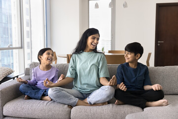 Cheerful Indian mom teaching yoga, training two kids at home. Young yogi mother and sibling...