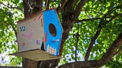 Small decorated birdhouse hanging from a tree branch. Painted with bright colours with children's stickers with sunlight in the leaves behind it
