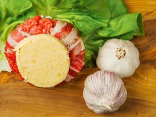 A beef burger wrapped in bacon or rasher and a slice of cheese on top of it and garlic cloves. The burger is surrounded by lettuce. The burger is on a wooden cutting board. High quality meat product