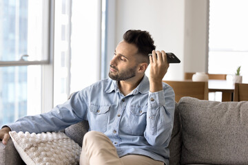 Fototapeta premium Serious young Latin man listening to voice message on mobile phone, sitting on cozy home sofa, looking away in deep thoughts, thinking on Internet communication, audio file