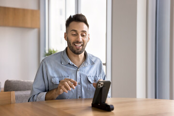 Fototapeta premium Happy Hispanic influencer man speaking at mobile phone web camera, giving business training, workshop, broadcasting on social media, blog on Internet, using phone on holder, talking on conference chat
